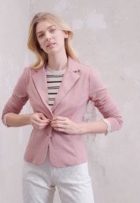 Blazer rose clair avec un col cranté, fermeture à boutons et manches longues, superposé à un pull rayé. Assorti avec un pantalon blanc à motifs.