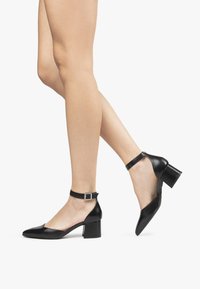 Scarpe pump in pelle nera con punta affusolata, cinturino alla caviglia e tacco a blocco. La texture liscia e il design minimalista accentuano l'eleganza.