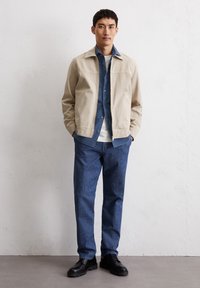 Beige canvas jacket over a blue button-up shirt, paired with dark blue denim pants and black lace-up shoes. Simple, structured design.