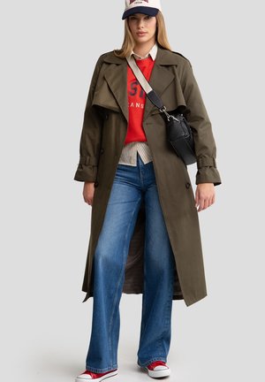 Trinchera color verde oliva, sudadera roja con texto, camisa a rayas, vaqueros de pierna ancha color azul y zapatillas rojas. Bolsa negra con correa.