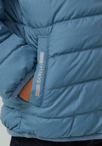 Hellblaue Steppjacke mit quiltierter Textur, seitlicher Tasche und horizontalen Nähten sowie reflektierendem Markenlabel in der Nähe der Tasche.