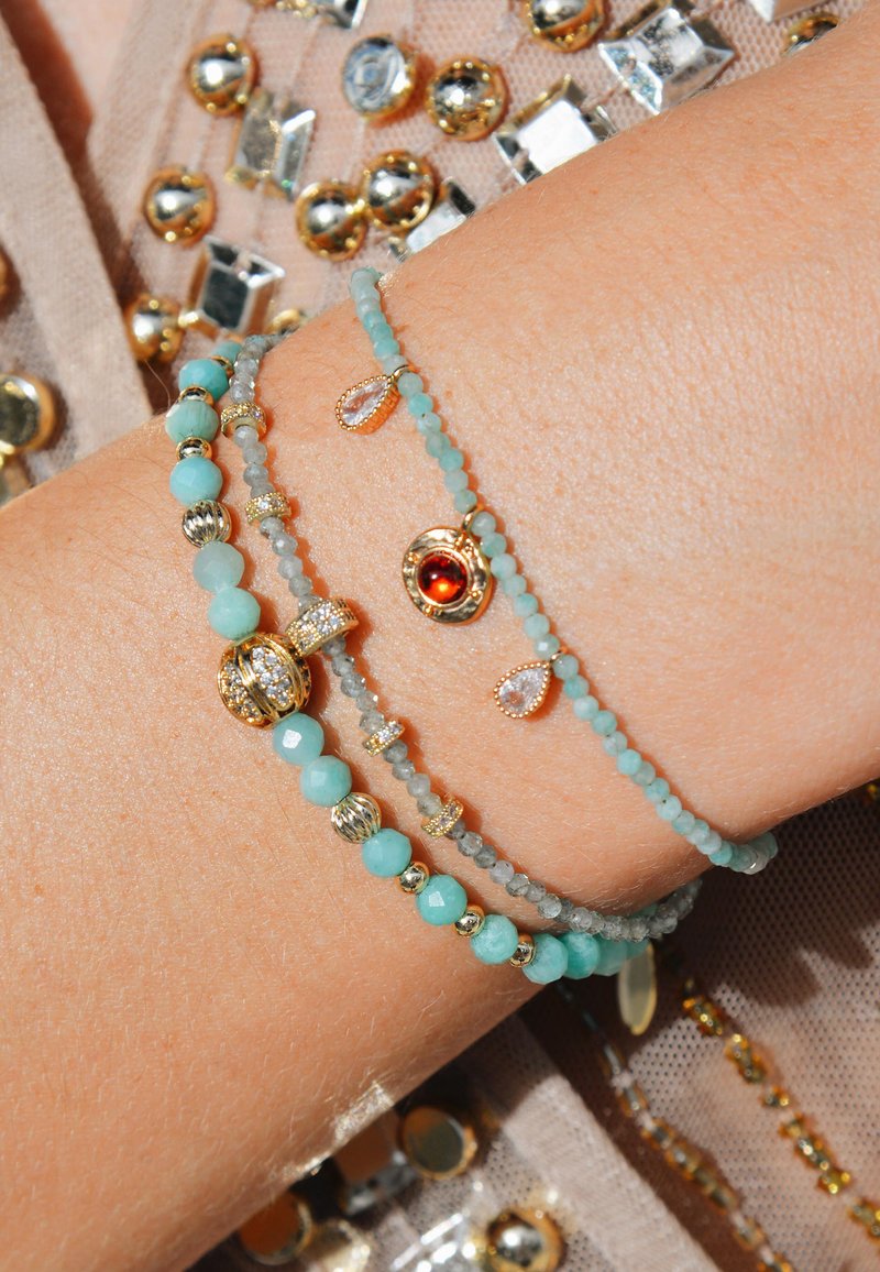 Trois bracelets de perles avec des accents turquoise, gris et dorés. L'un d'eux met en avant une pierre précieuse rouge et de petits charms en forme de goutte. Les perles varient en taille et en texture.