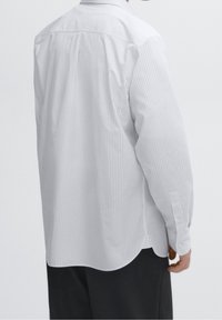 Chemise blanche à manches longues avec fines rayures verticales, ourlet incurvé et poignets boutonnés. Le tissu semble léger et lisse. Vue de dos.