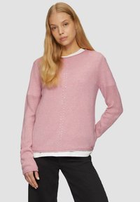 Lichtroze gebreide trui met ribbelstructuur, ronde halslijn, lange mouwen en subtiele verticale details aan de voorkant. Gedragen over een wit shirt.