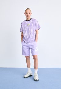 Violeta krāsu t-krekls ar dzeltenu logo, kombinēts ar gaiši violetām šortiem, kuros ir balta Nike logo, un baltām zeķēm ar pelēkiem un melniem apaviem.