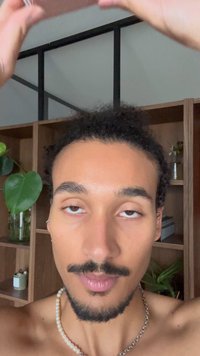 Ung mand med krøllet hår og skæg løfter hænderne, stående foran trähylder med planter og dekorationsgenstande.