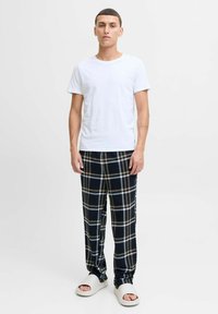 T-shirt bianca in cotone, pantaloni di flanella a quadri neri e beige, e ciabatte in gomma bianche, in piedi su uno sfondo neutro.
