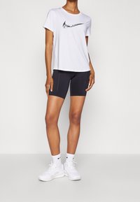 Camiseta blanca de Nike con logo negro, combinada con pantalones cortos de ciclismo negros. El modelo lleva zapatillas blancas y calcetines, mostrando un look atlético casual.