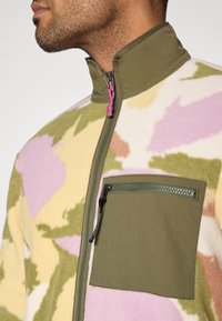 Ζακέτα από φλις με καμουφλάζ μοτίβο σε ροζ, πράσινο και κίτρινο. Διαθέτει ψηλό γιακά και τσέπη με φερμου άρ στο στήθος.