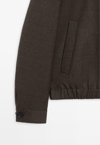 Manica di giacca in tessuto marrone scuro con polsino con bottoni, tasca laterale con spacco e dettaglio orlo elasticizzato su sfondo bianco.