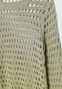 Maglione lavorato a maglia verde chiaro con un design a traforo che presenta grandi fori. Collo rotondo e vestibilità ampia con filato morbido e strutturato.