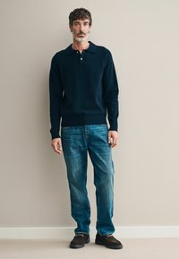 Tengerészkék kötött pulóver rövid gallérral, kék farmernadrág és sötétbarna slip-on cipő, gumitalppal, egy bézs fal előtt állva.