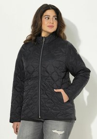 Kvinna iklädd en svart quiltad zip-up jacka med händerna i fickorna, i sällskap med grå slitna jeans, stående mot en enkel ljus bakgrund.