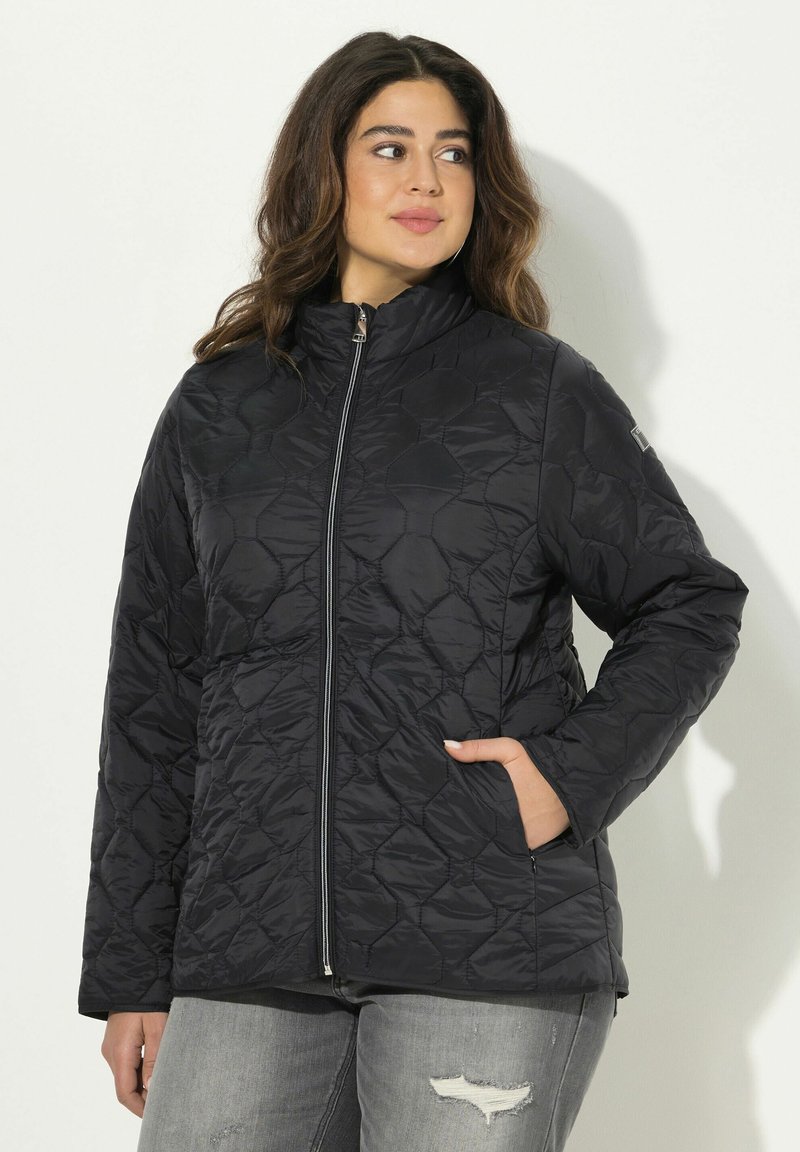 Kvinna iklädd en svart quiltad zip-up jacka med händerna i fickorna, i sällskap med grå slitna jeans, stående mot en enkel ljus bakgrund.