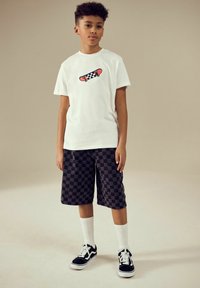 Hvid t-shirt med et farverigt skateboardgrafik, parret med sorte ternede shorts og sorte sneakers, suppleret med hvide sokker.