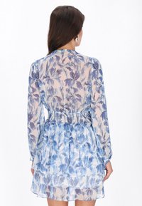 El vestido floral presenta una tela translúcida azul y blanca, mangas largas, cintura fruncida y un dobladillo con volantes en capas con una textura ligera.