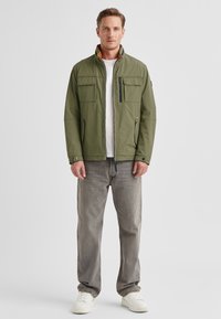 Olive green jacket with a high collar, front zipper, and two side pockets. Worn over a white shirt, paired with gray jeans and white sneakers.