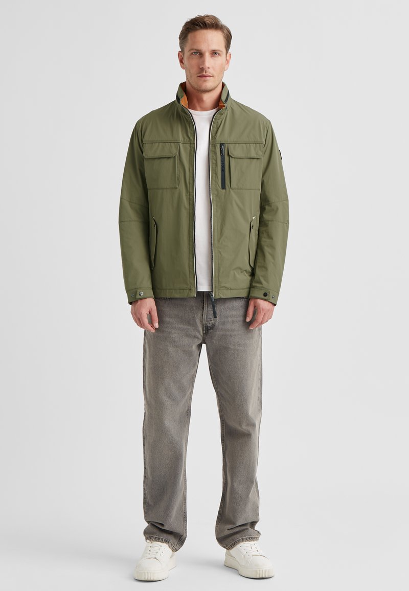 Olive green jacket with a high collar, front zipper, and two side pockets. Worn over a white shirt, paired with gray jeans and white sneakers.