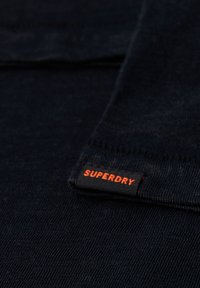 Felpa di un tessuto navy con una trama a coste, dotata di un'etichetta nera con la scritta "SUPERDRY" in arancione brillante. Design semplice e pulito.