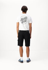 Camiseta blanca con el logotipo negro de "Santa Cruz" en la espalda, combinada con pantalones cortos de cargo negros, terminando con zapatillas azules y calcetines blancos de caña corta.