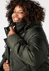 Gevoerde pufferjas in donkergroen, met een hoge kraag, frontale rits en aantrekdetails. Effen textuur met lichte isolatie.