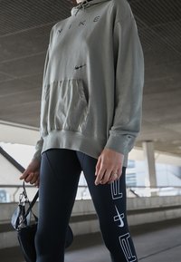 Světle olivová mikina s kapucí a přední kapsou ve stylu klokanky, doplněná o černé legíny s bílým grafickým potiskem a detailem na zip.