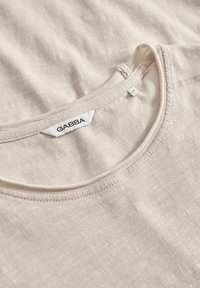 Beigā kokvilnas T-krekls ar apaļu kakla izgriezumu un smalku tekstūru. Iekšējā apkaklē atrodas GABBA etiķete. Tīra, vienkārša šuvšana pa malām.