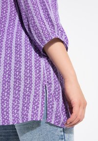 Lilla bluse med stjernemønster, opsmøgede ærmer, sideslids og en struktureret stof, parret med lyseblå jeans.