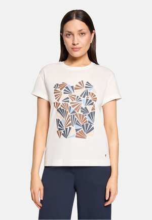 Woman with straight dark hair wearing a white short-sleeve t-shirt with geometric brown, blue, and beige fan patterns and dark pants.