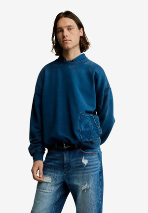 Giovane uomo con capelli fino alle spalle che indossa una felpa blu con una toppa e jeans blu consumati, in piedi contro uno sfondo bianco.