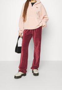 Överdimensionerad rosa fleecehoddie med halvmiss, i kombination med vinröda corduroybyxor och kraftiga vita skor. En svart handväska syns också.