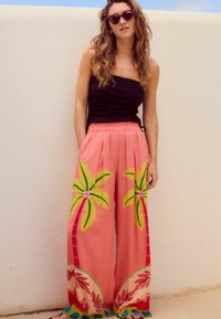 Pantalons larges roses avec imprimés de palmiers en vert et jaune, dotés d'une taille élastique. Associé à un haut noir et des lunettes de soleil rondes.