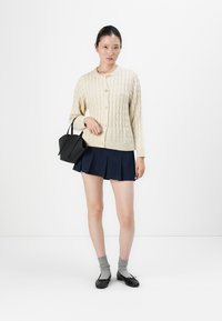 Ung kvinne iført en kremfarget kabelstrikket cardigan, marineblå plisserte skjørt, grå sokker, svarte leiligheter, holder en liten svart håndveske mot hvit bakgrunn.