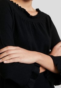 Personne portant un haut noir à manches longues avec col et poignets à volants, bras croisés sur la poitrine, fond neutre.