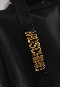 Sangle de sac en tissu noir avec des lettres dorées brillantes épelant "MOSCHINO" attachées verticalement.