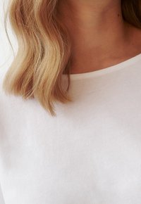 T-shirt bianca in cotone con scollo rotondo, morbido al tatto e maniche corte. I capelli appaiono ondulati e biondi dorati, incorniciando l'area del collo.