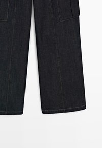 Pantalon en jean indigo foncé à jambes larges avec coutures apparentes et poches avant rectangulaires sur fond blanc.