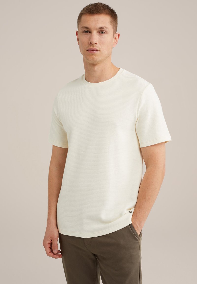 Tricou scurt cu mânecă scurtă de culoare crem deschis, cu un model texturat, guler rotund și un logo mic accentuat la tiv. Asortat cu pantaloni de culoare verde măslinie.
