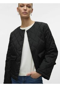 Juodasis quiltuotas džemperis su apvaliu kaklu, sagų uždarymu, krūtinės kišeniu ir tekstūruotu audiniu. Dėvimas ant baltos marškinėlės ir džinsų.