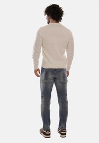Uomo con capelli ricci che indossa un maglione beige, jeans blu sbiaditi con macchie di vernice e sneakers Nike marroni, in piedi di spalle.