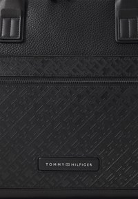 Black leather bag with textured surface, featuring a geometric pattern. Secure zipper closure and logo tag on the front for branding.