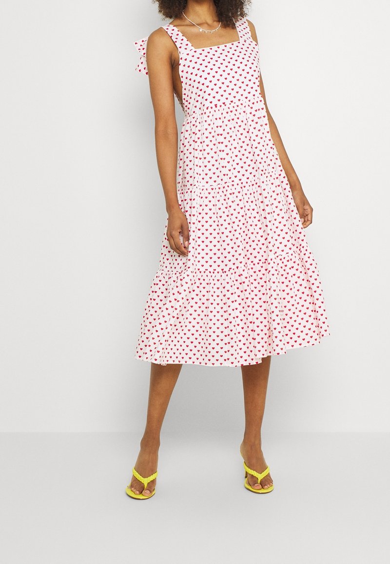 Robe sans manches blanche avec un motif de cœurs rouges, décolleté carré et jupe évasée. Présente des détails de nœud aux épaules et un ourlet à volants superposés.