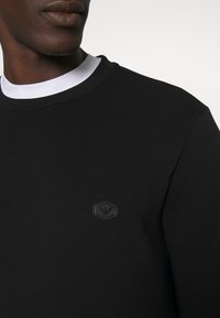 Man med svart rundhalsad sweatshirt och vit undertröja, med en liten hexagonal logotyp på vänster bröst.
