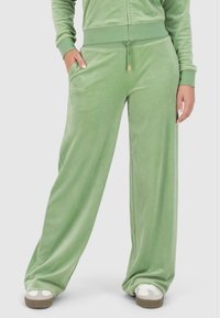 Pantalons en velours vert à coupe loose, avec une taille élastique, des poches latérales et une texture lisse. Présente des accents en métal doré.