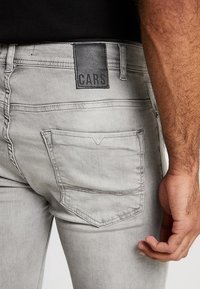 Primer plano de una persona con jeans gris claro que tiene un bolsillo trasero y una etiqueta negra con la palabra "CARS" en la cinturilla.