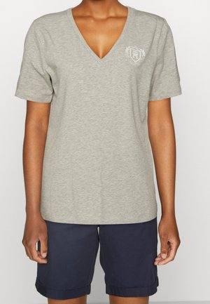 Persoon die een grijze V-hals T-shirt met korte mouwen draagt met een klein wit embleem en marineblauwe opgerolde shorts, staand tegen een effen achtergrond.