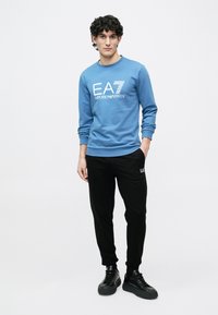 Μπλε φούτερ με λευκό τυπωμένο λογότυπο "EA7", συνδυασμένο με μαύρη φόρμα με λεπτομέρεια λογότυπου και μαύρα αθλητικά παπούτσια. Ομαλή υφή υφάσματος.