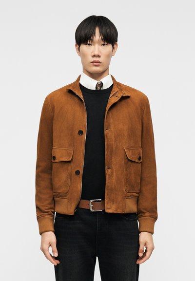 Homme portant une veste en daim marron sur un pull noir et une chemise blanche avec une cravate à motifs, se tenant devant un fond gris uni.