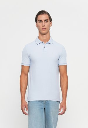 Young man with short slicked-back hair wearing a light blue short-sleeve polo shirt and light blue jeans, standing against a white background.