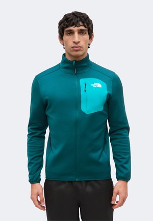 Hombre con chaqueta de cremallera completa color verde azulado con bolsillo en el pecho con cremallera de color verde azulado más claro y logotipo negro sobre fondo claro y liso.